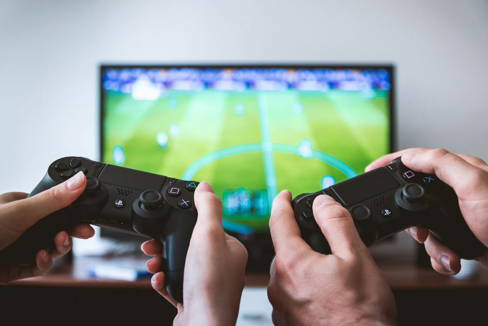 Two sets of hands holding PS5 controllers, playing a game on an out-of-focus TV.